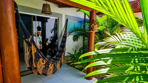 A hammock hanging between wooden pillars in a covered outdoor area with a view of a building and lush green plants. - Chalé Recanto dos Passarinhos  (Tibau do Sul)