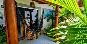 A hammock hanging between wooden pillars in a covered outdoor area with a view of a building and lush green plants. - Chalé Recanto dos Passarinhos  (Tibau do Sul)