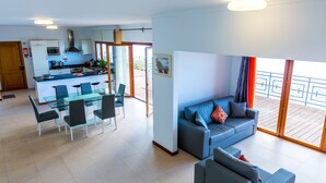 Ein modernes Wohnzimmer mit grauem Sofa, einem Glastisch und Stühlen. Es gibt eine Küchenzeile mit Mikrowelle und Herd, sowie einen Balkon mit Geländer.