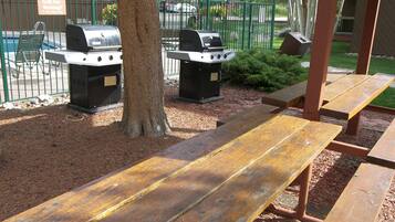 A picnic area with two barbecue grills, a wooden bench, and a fenced playground in the background.