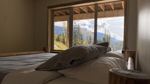 Een slaapkamer met een groot raam, houten balken, een bed met wit beddengoed en een houten nachtkastje.