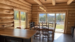 Het interieur van een blokhut met een houten eettafel en stoelen, een keuken met een vaatwasser, en grote ramen met uitzicht op het bos en de bergen.