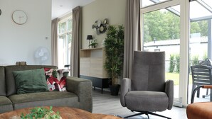 Una sala de estar moderna con sofá, sillón y mesa de centro. Hay un ventanal con cortinas, un ventilador y una planta en maceta.
