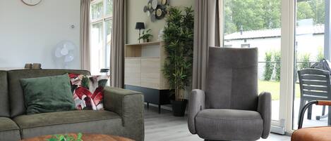 Een moderne woonkamer met een bank, fauteuil en salontafel. Er is een groot raam met gordijnen, een ventilator en een potplant.