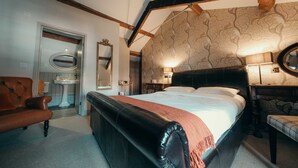 A bedroom with a large bed, a leather bench, a wooden ceiling, and a bathroom with a sink and mirror.