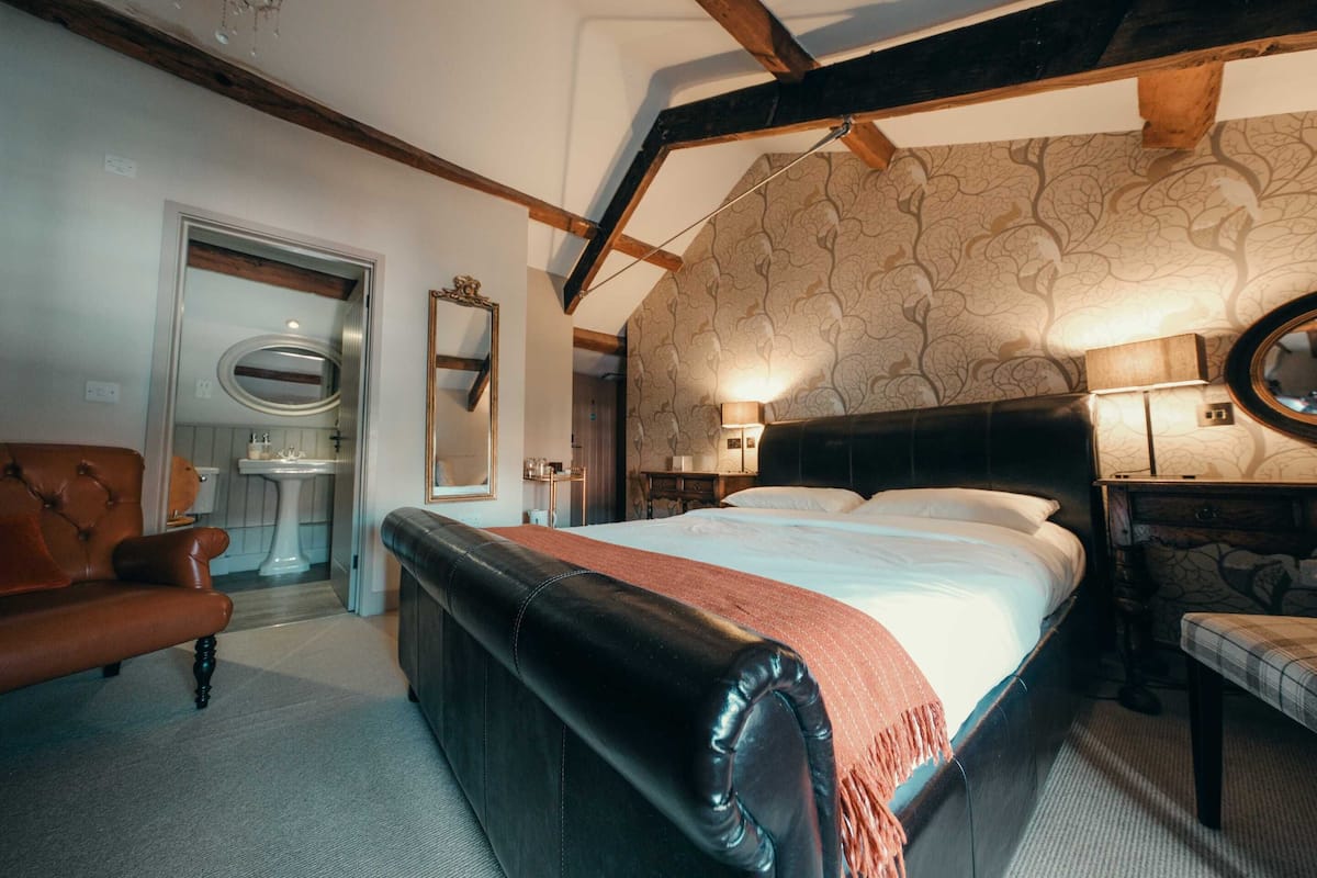 A bedroom with a large bed, a leather bench, a wooden ceiling, and a bathroom with a sink and mirror.