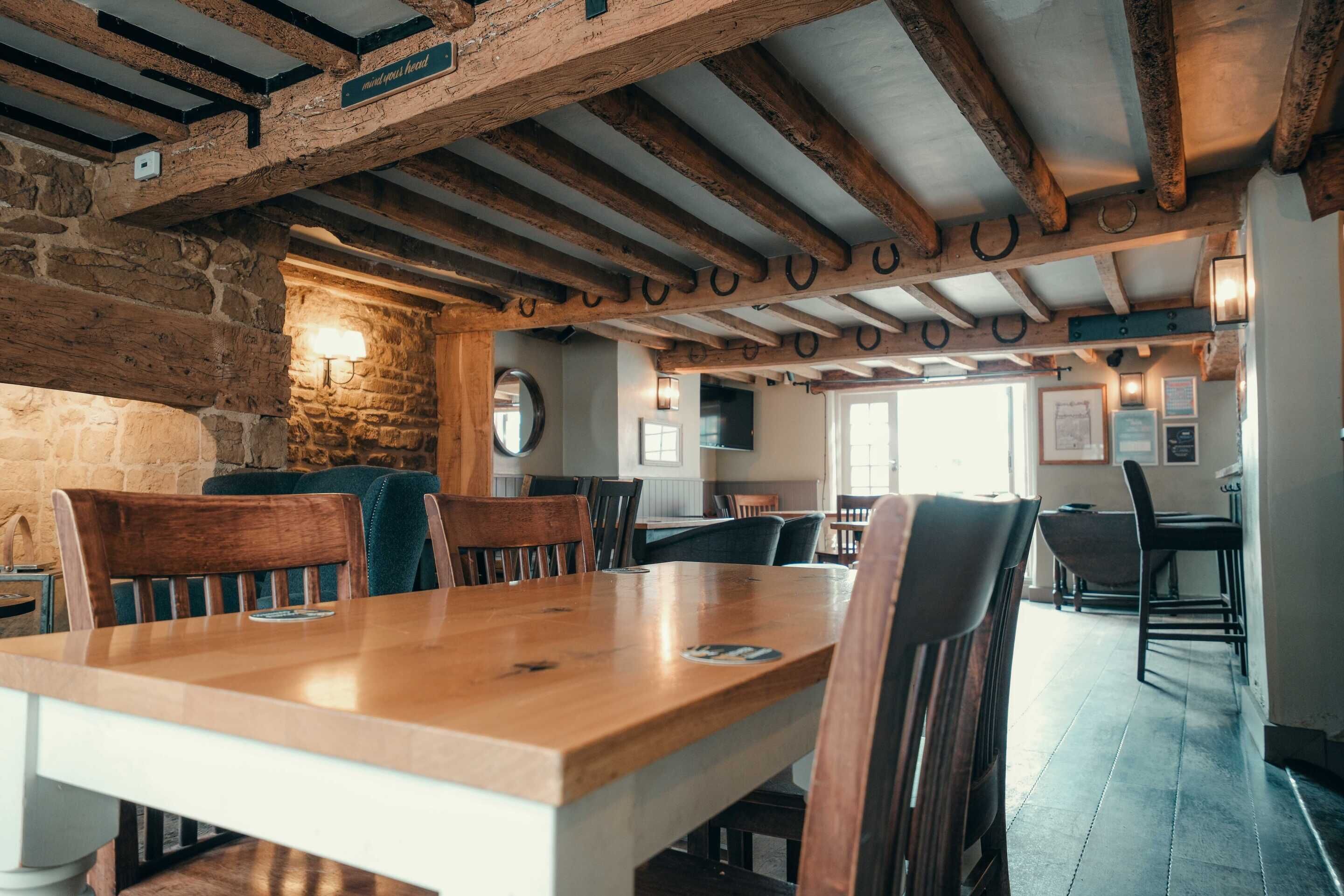 A rustic interior with exposed wooden beams, stone walls, and a long wooden table with chairs.
