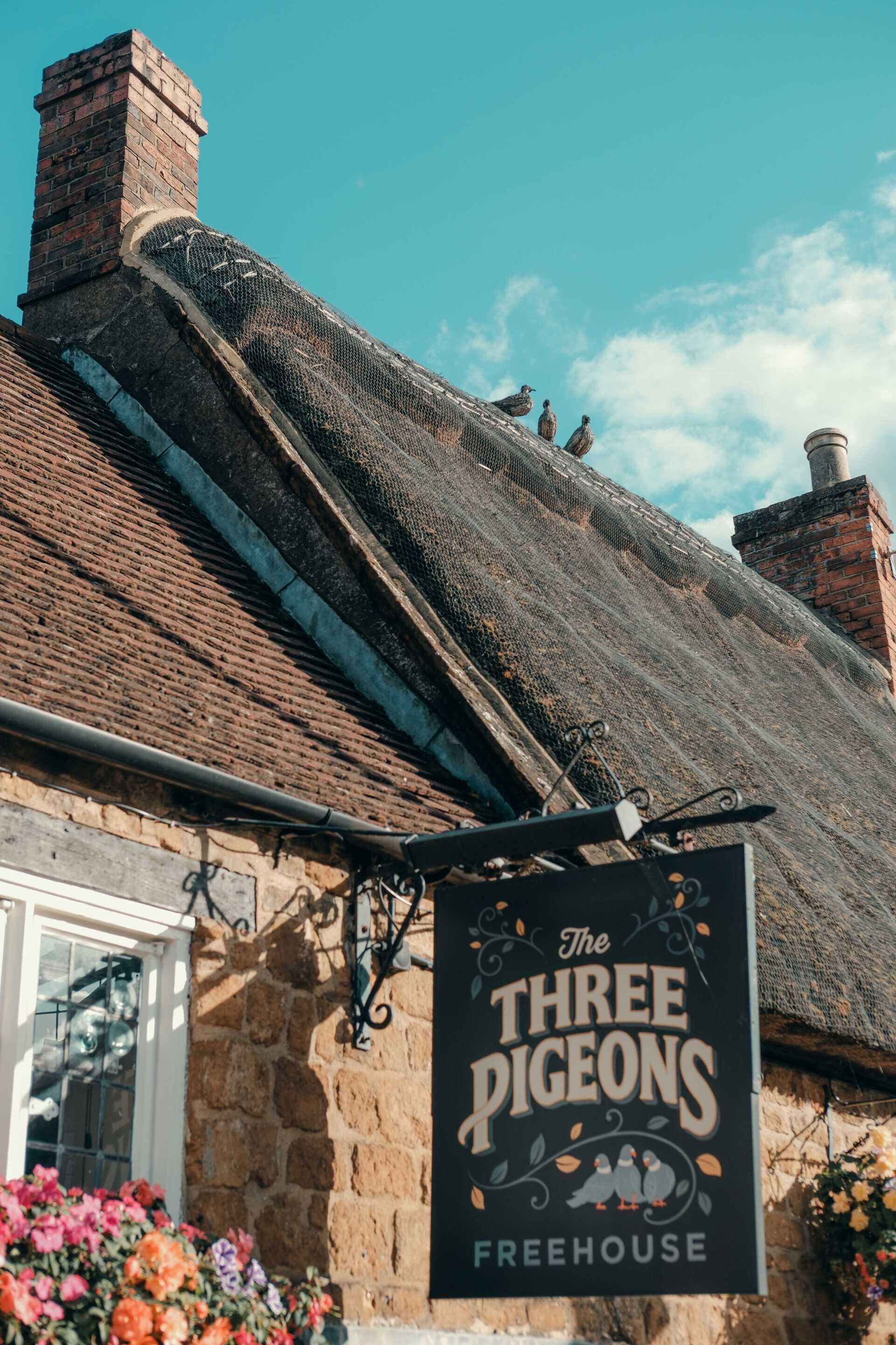 A building with a sign reading 'The Three Pigeons Freehouse,' a stone facade, and a thatched roof.