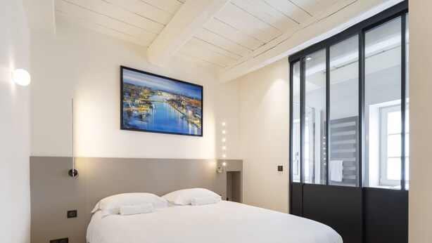 Een moderne slaapkamer met een groot bed, een stadsgezicht schilderij erboven, een spiegel met verlichting en een schuifglazen deur aan de rechterkant.