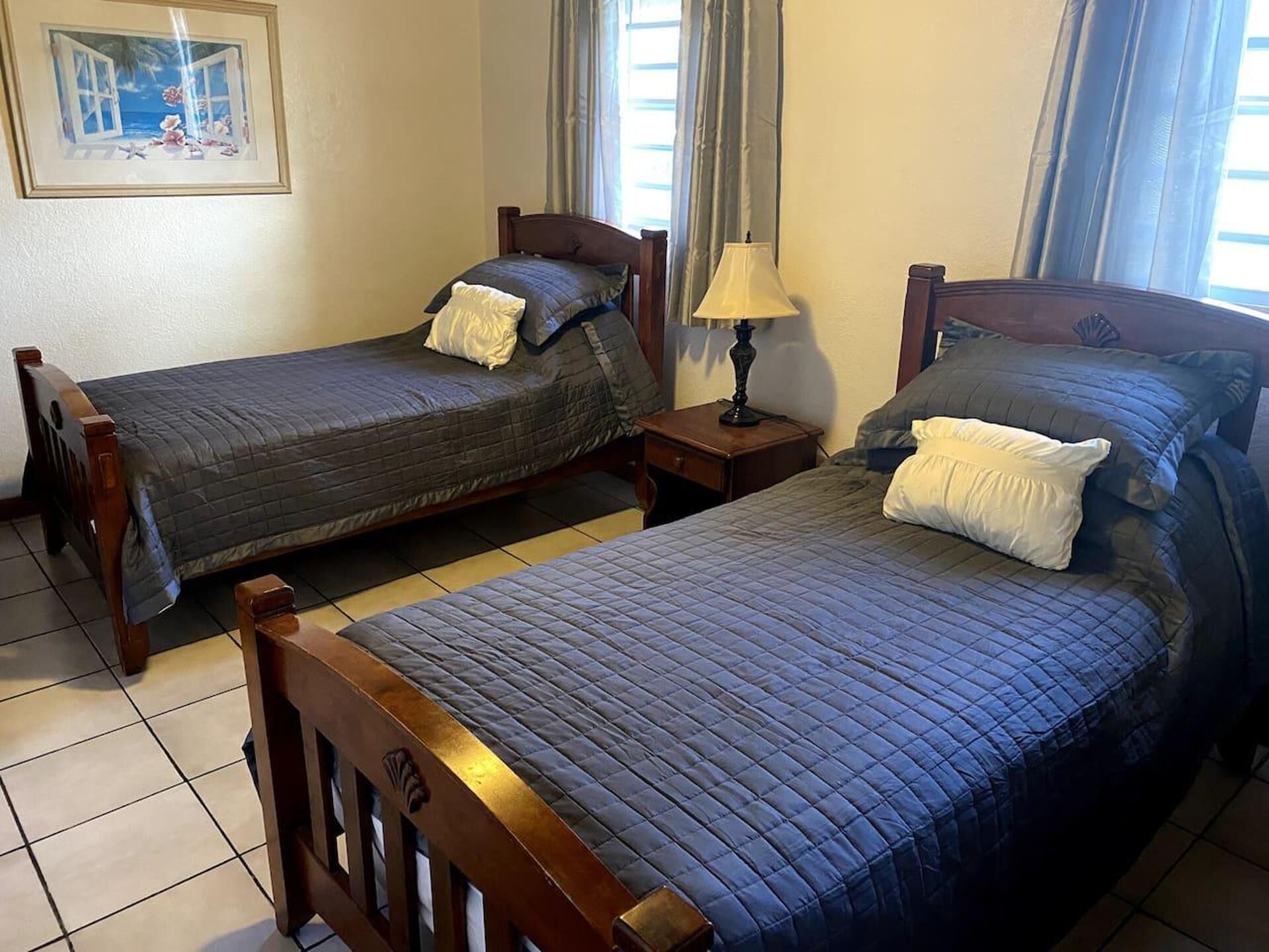 A room with two wooden beds, each with a quilted blue bedspread and pillows. There is a small wooden nightstand with a lamp between the beds. A framed picture hangs on the wall above the left bed.