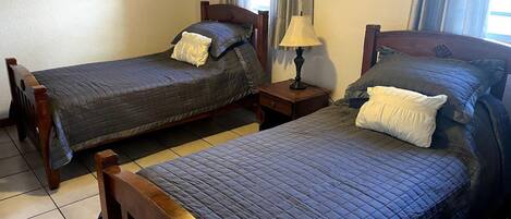 A room with two wooden beds, each with a quilted blue bedspread and pillows. There is a small wooden nightstand with a lamp between the beds. A framed picture hangs on the wall above the left bed.