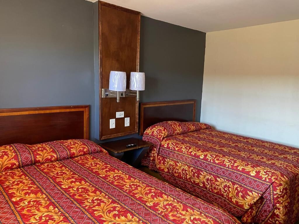 A hotel room with two beds, each with a red and gold patterned bedspread. There is a wooden wardrobe with a mirror and two wall-mounted lamps.