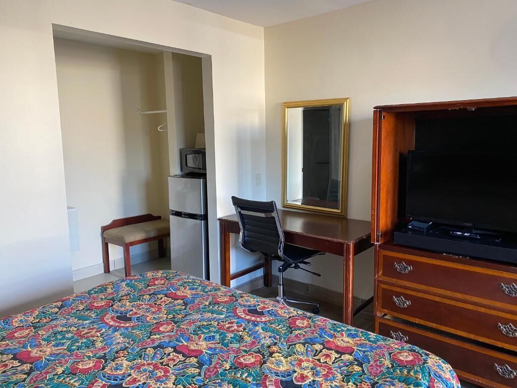 Quarto de hotel com cama, escrivaninha, cadeira, televisão e guarda-roupa.