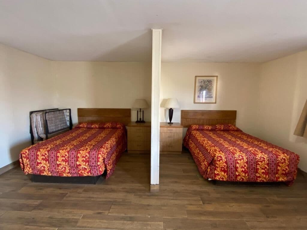 Una habitación con dos camas individuales, cabeceros de madera, una mesita de noche con lámpara y un cuadro enmarcado en la pared.