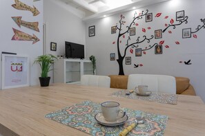A modern dining area with a wooden table, two chairs, a television, and a wall decorated with a tree and pictures. - Tana del Bianconiglio by Wonderful Italy (Turin)
