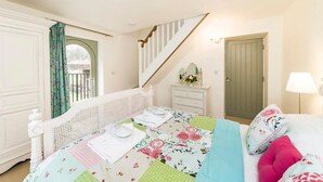 Ein Schlafzimmer mit Bett, Kommode, einem Fenster mit Vorhängen, einer Tür und einer Treppe.