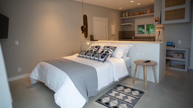 A modern bedroom with a large bed, bedside tables, a small stool, and built-in shelving with decorative items. - Koserê (Ilhabela)