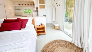 A modern bedroom with a large bed, a small wooden stool, a woven basket, and a view of the outdoors through a large window. - Koserê (Ilhabela)