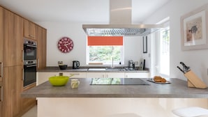 A modern kitchen with wooden cabinets, a large window, and a bright red clock. - Stylish 4 bed house walking distance to beaches (Salcombe)