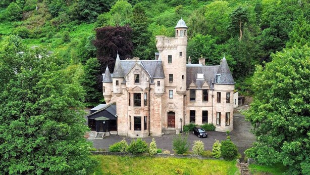 Un grand bâtiment historique en pierre, doté de nombreuses tours et d’une entrée centrale, le tout entouré d’une végétation luxuriante.