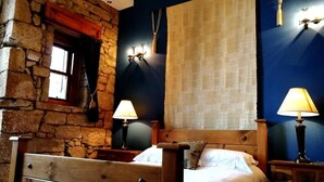 Une chambre avec un mur en pierre, un lit en bois, deux tables de chevet avec des lampes et une grande fenêtre avec des rideaux.