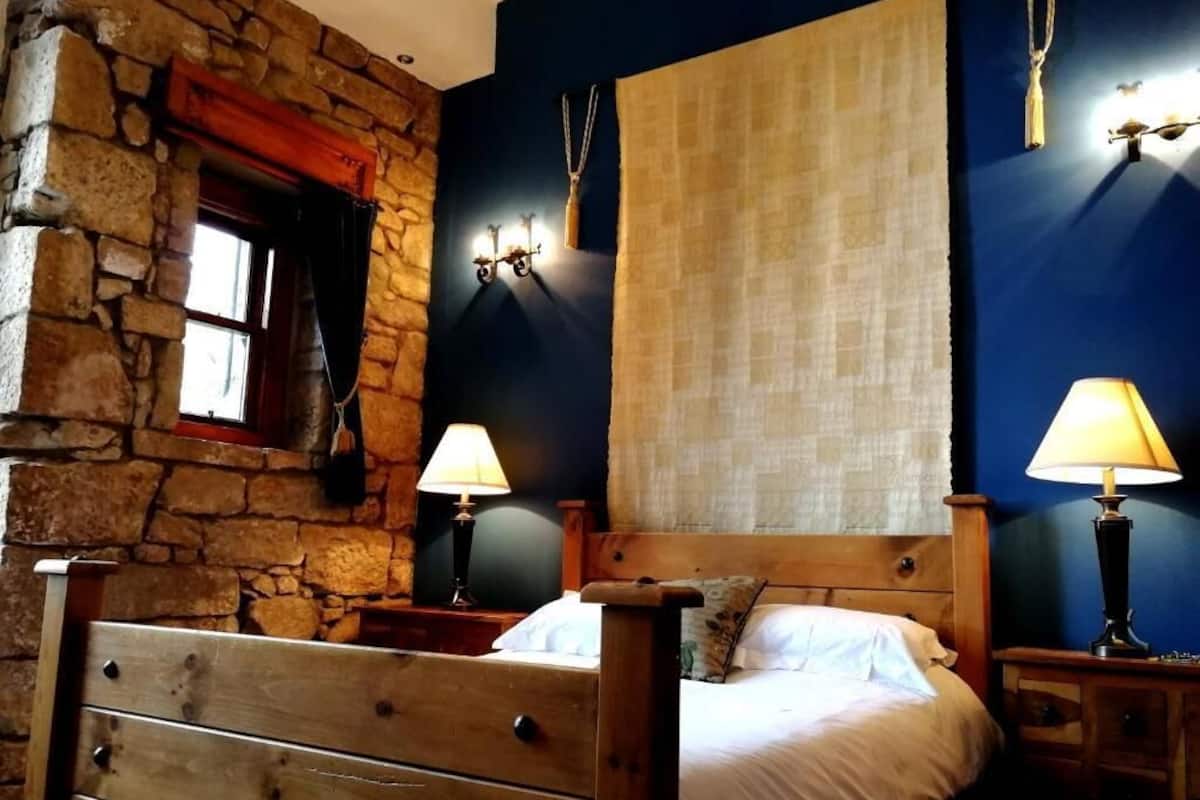 A bedroom with a stone wall, a wooden bed, two bedside tables with lamps, and a large window with curtains.