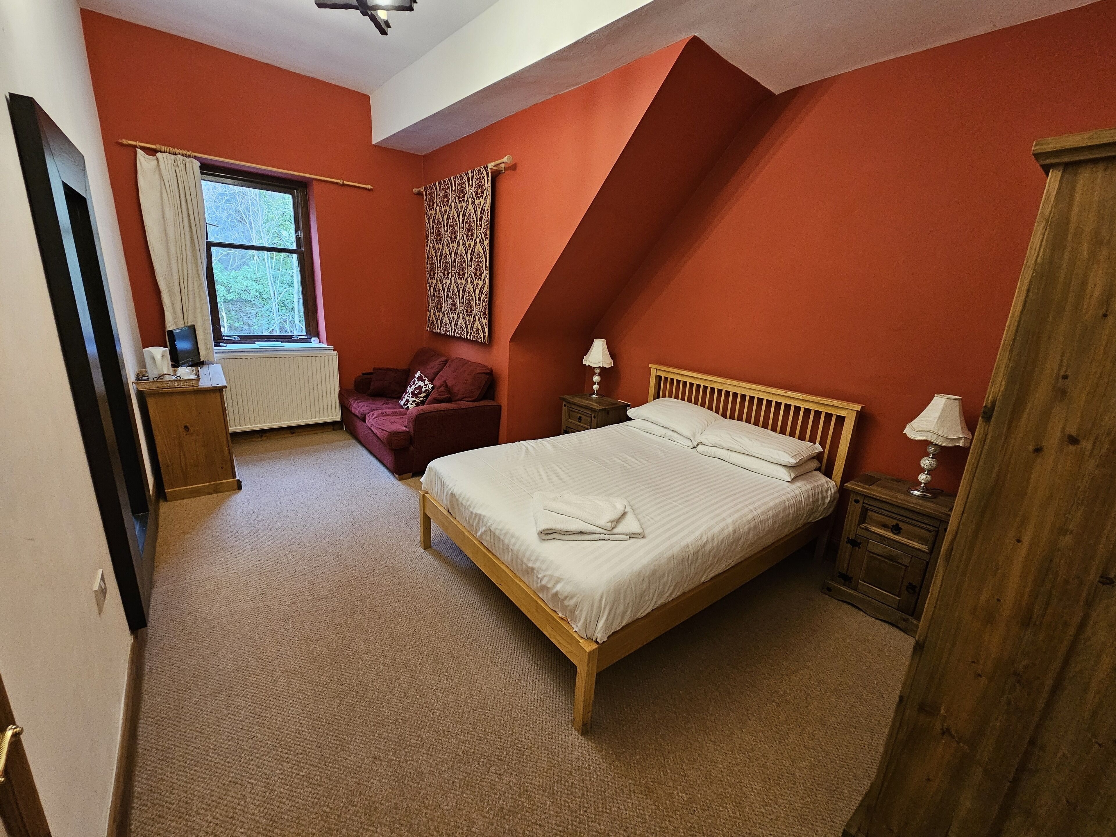 A bedroom with a bed, a window, a red wall, a wooden wardrobe, and a small table with a lamp.