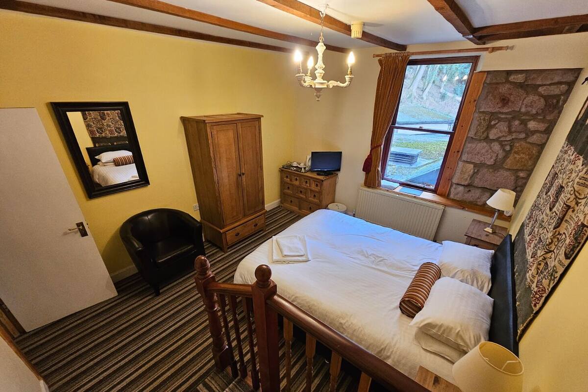 A bedroom with a bed, a wooden wardrobe, a desk with a computer, a chair, a mirror, and a window with a view of the outdoors.