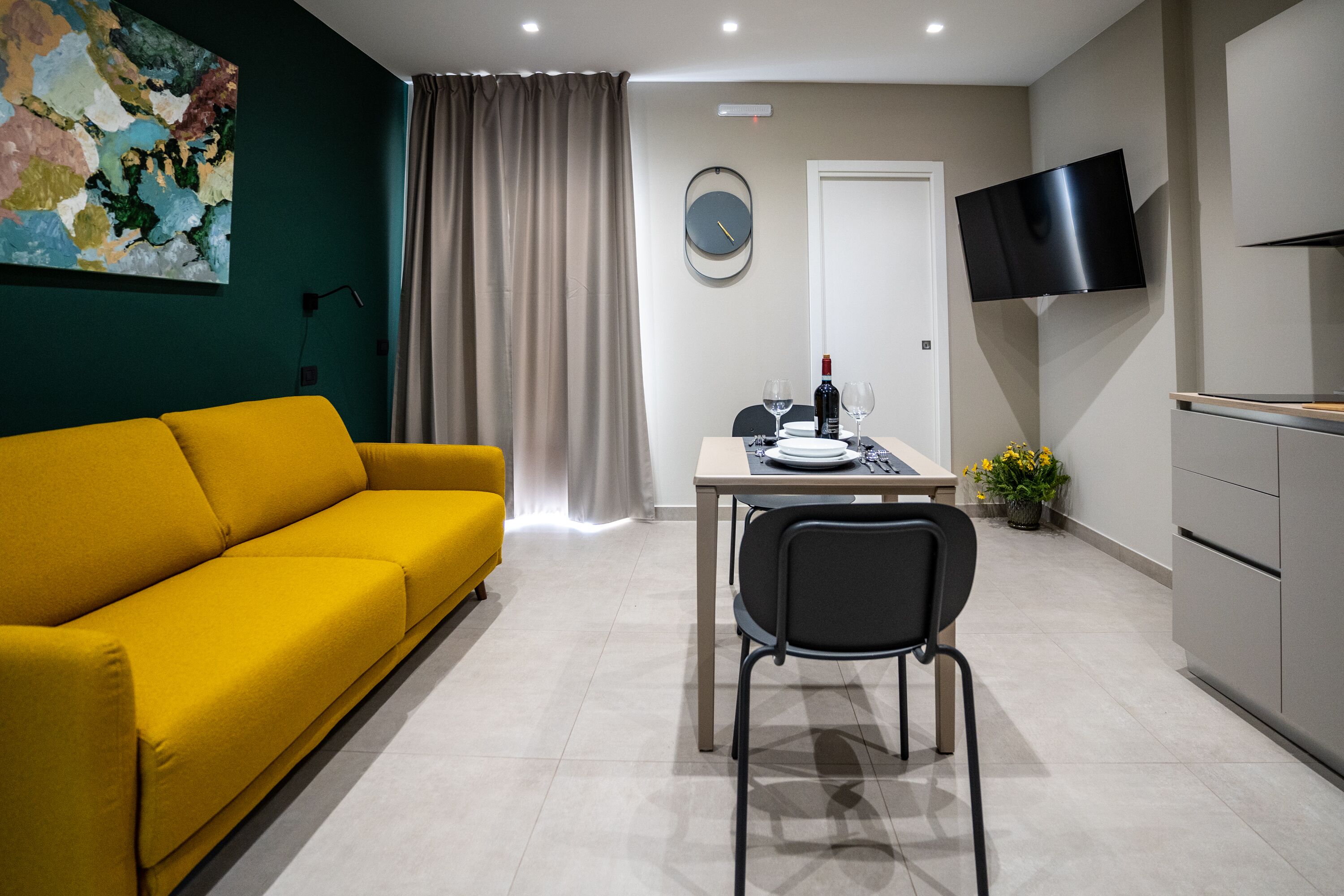Uma sala de estar moderna com um sofá amarelo, uma mesa de jantar com cadeiras, uma televisão de tela plana e um relógio na parede.