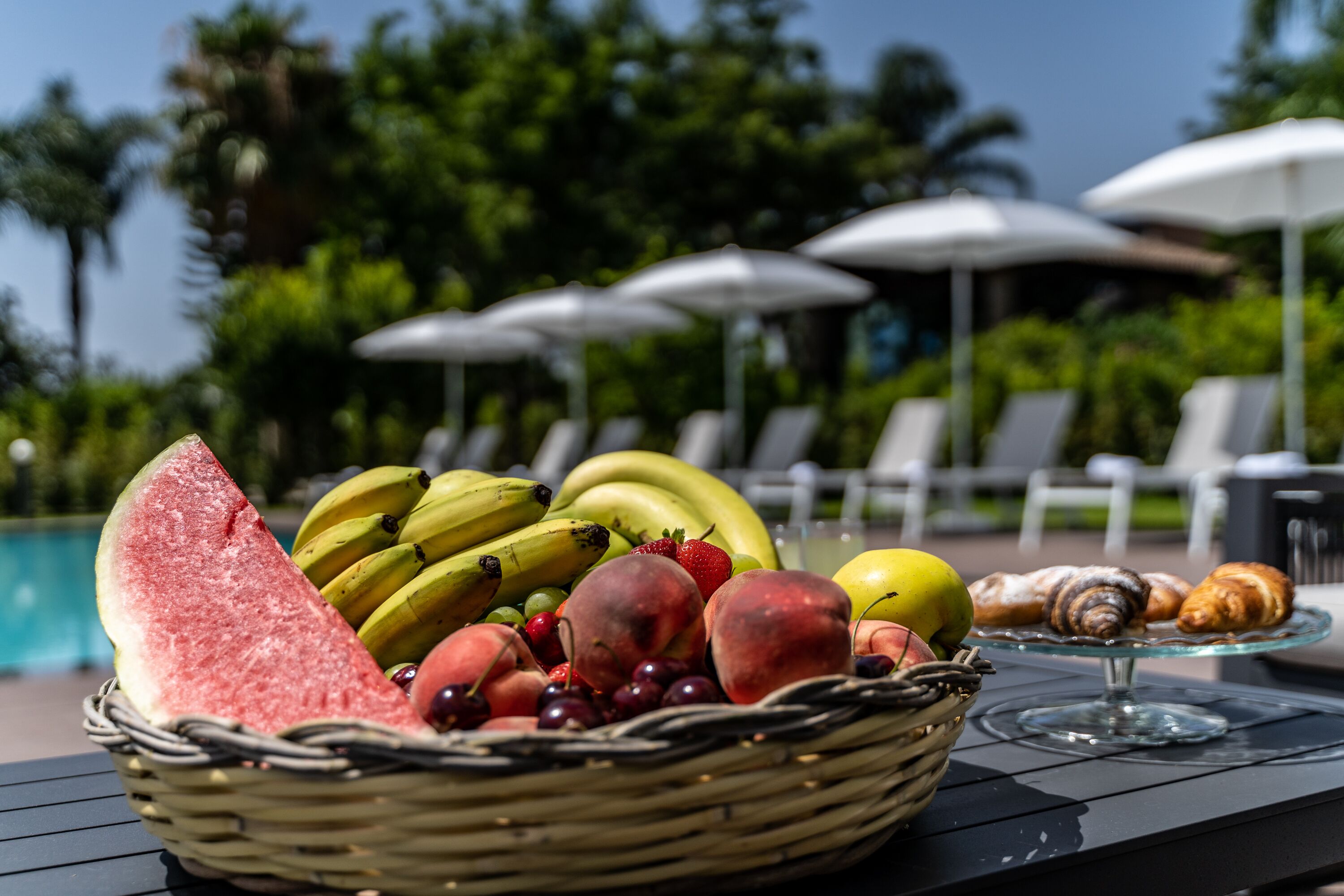 Una cesta de frutas con sandía, plátanos, uvas y melocotones sobre una mesa junto a una piscina, con sillones de descanso y sombrillas al fondo.