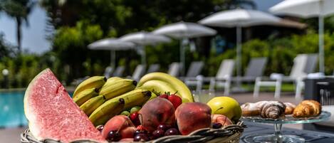 Una cesta de frutas con sandía, plátanos, uvas y melocotones sobre una mesa junto a una piscina, con sillones de descanso y sombrillas al fondo.