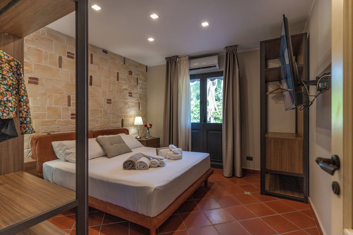 Ein Hotelzimmer mit einem Bett, einem hölzernen Kopfteil, einem Nachttisch mit Lampe, einem Kleiderschrank und Blick ins Freie durch eine Tür.