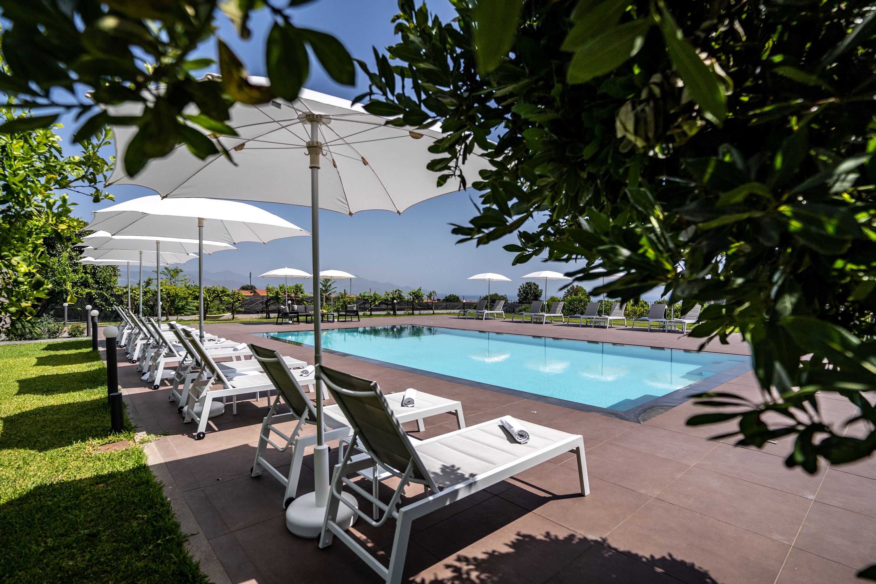 Un espace aménagé au bord de la piscine, avec des chaises longues blanches et des parasols, une piscine à l’eau d’un bleu limpide, et une vue sur les montagnes en arrière-plan.