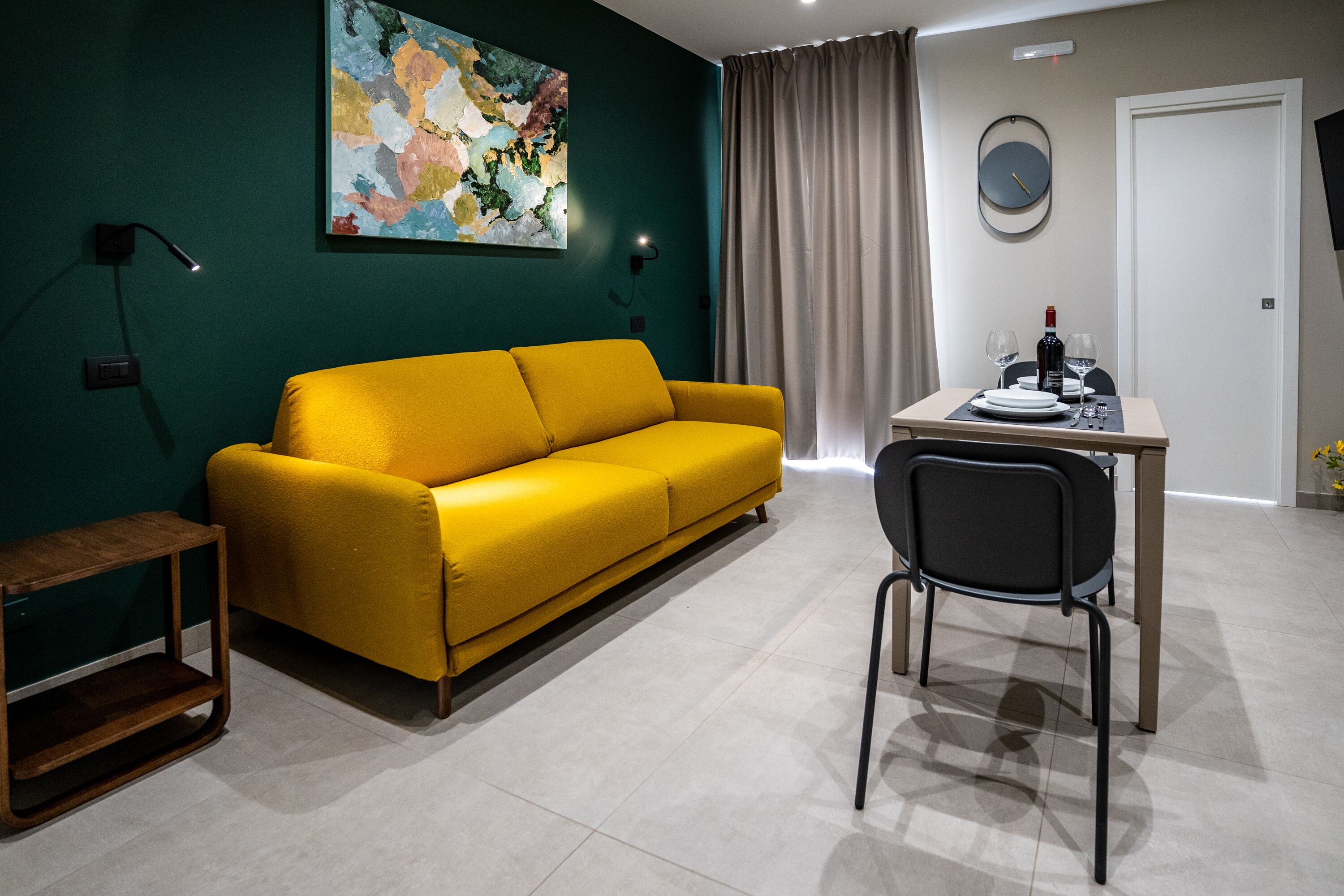 A modern living room with a yellow sofa, a dining table with two chairs, a small side table, a wall-mounted lamp, a painting, a clock, and a door.