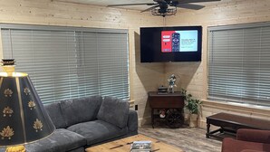 Ein Wohnzimmer mit einer grauen Eckcouch, einem Holztisch, einem an der Wand montierten Fernseher und einem Deckenventilator.