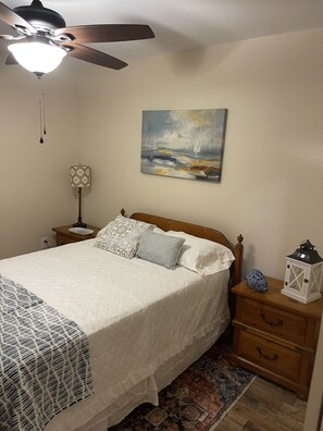 Un dormitorio con cama, mesitas de noche, ventilador de techo, un cuadro y una luz nocturna.