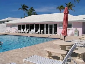 A swimming pool with lounge chairs and umbrellas, surrounded by palm trees and a building with a pink facade.