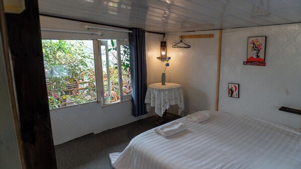 A bedroom with a bed, a small table with a vase, a hanging light, and a window with curtains.