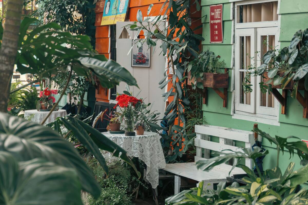 Un jardín con una mesa blanca, plantas en macetas y un edificio verde con un cartel.