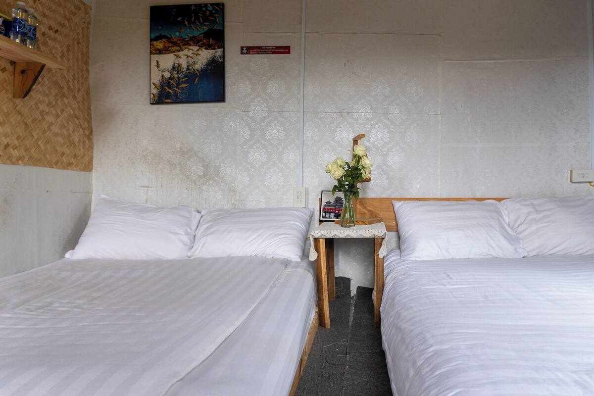 Una habitación de hotel con dos camas individuales, una mesita de noche de madera con un jarrón de flores, un cuadro en la pared y un estante para botellas de agua.