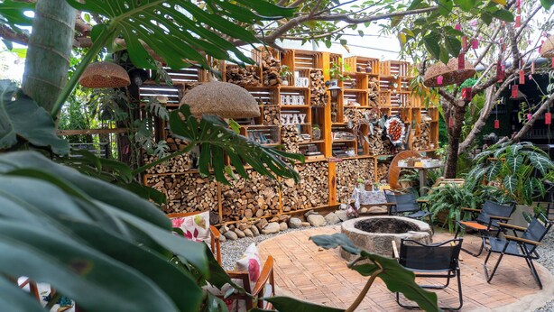 A rustic outdoor seating area with wooden furniture and a fire pit, surrounded by wooden shelves filled with plants and decorative items. - Gio Homestay  (Xuan Huong - Da Lat)