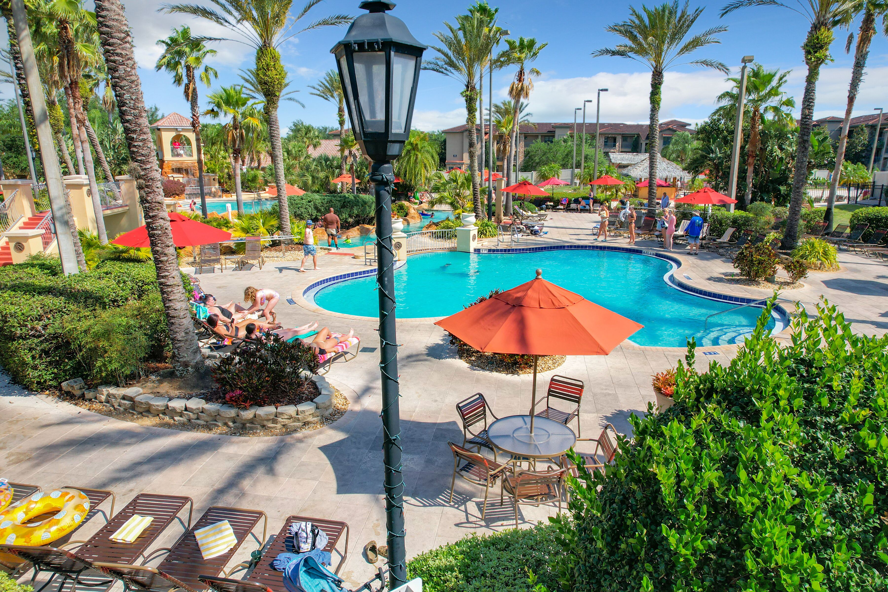 A resort pool area with a large pool, lounge chairs, umbrellas, and palm trees.