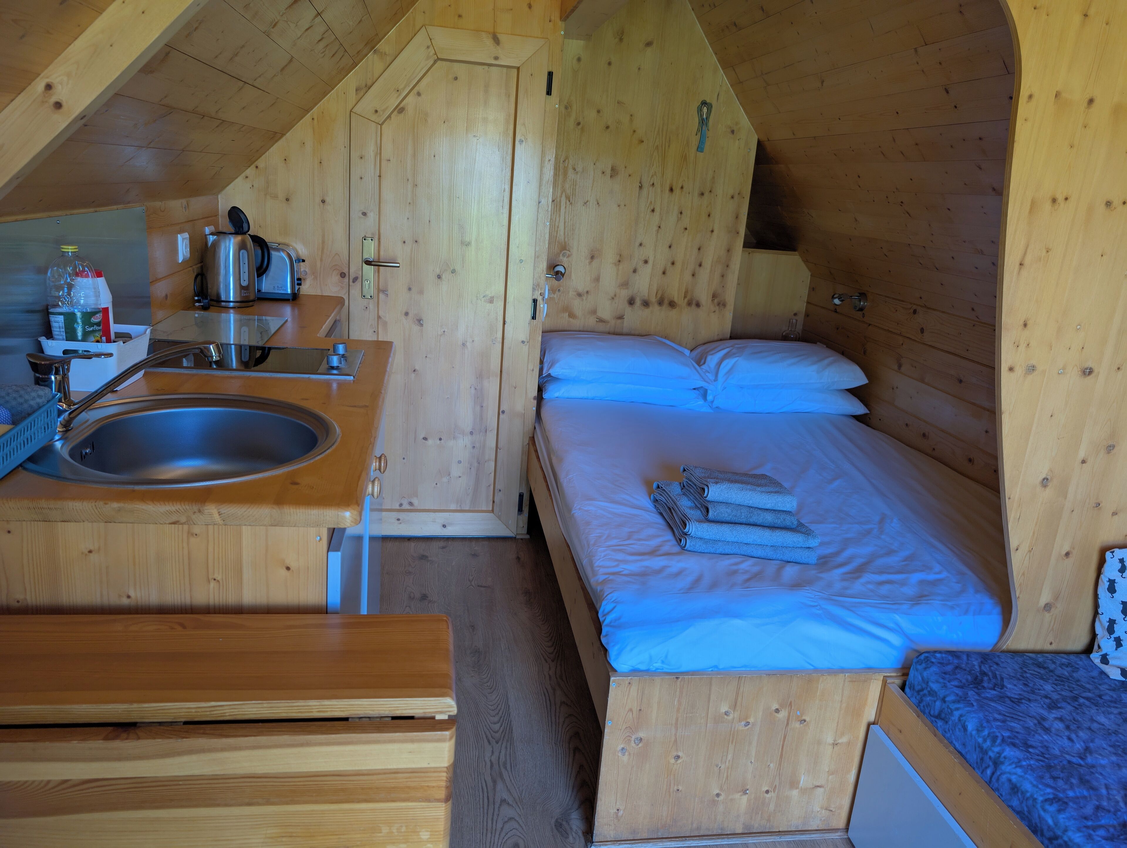 Una cabaña de madera compacta con kitchenette, una cama con ropa de cama azul y una pequeña mesa.