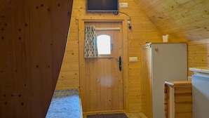 Une pièce chaleureuse, aménagée dans une cabane en bois, avec un lit, un petit bureau, une télévision et une porte dotée d’une fenêtre.