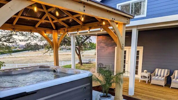 Un bain à remous sous une pergola en bois, avec vue sur une maison et sur du mobilier d’extérieur.