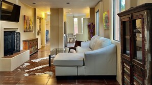 A spacious living room with a white sofa, a glass coffee table, and a fireplace. There is a dining area with a table and chairs in the background. - Charming Adobe Casita! Large lot in upscale quiet neighborhood. STR 15734641 (Fountain Hills)