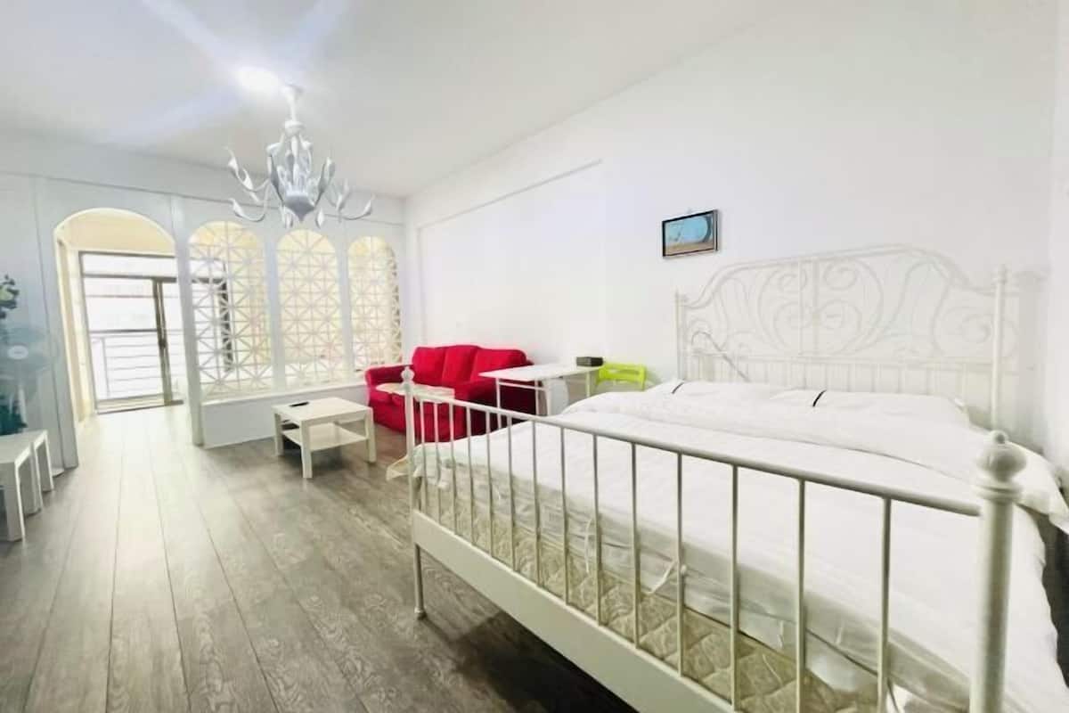 Ein Schlafzimmer mit weißem Metallbettgestell, einem roten Sessel, einem kleinen Tisch und einem Fenster mit geometrischem Muster.