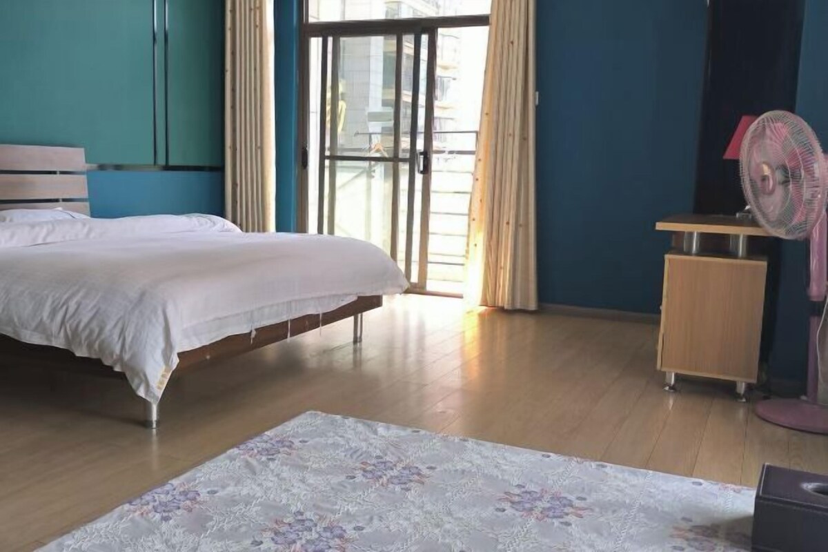A bedroom with a bed, a wooden desk, a standing fan, and a window with curtains.