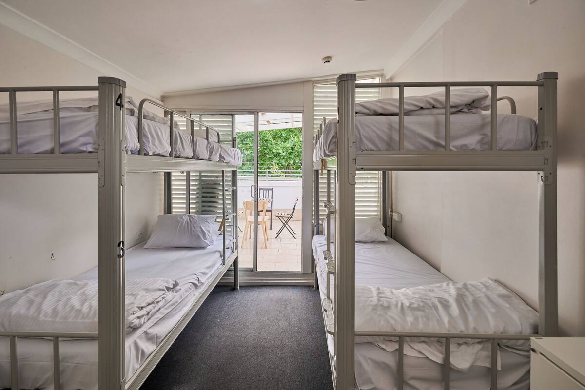 A dormitory room with bunk beds, a small table, and chairs near a sliding glass door.