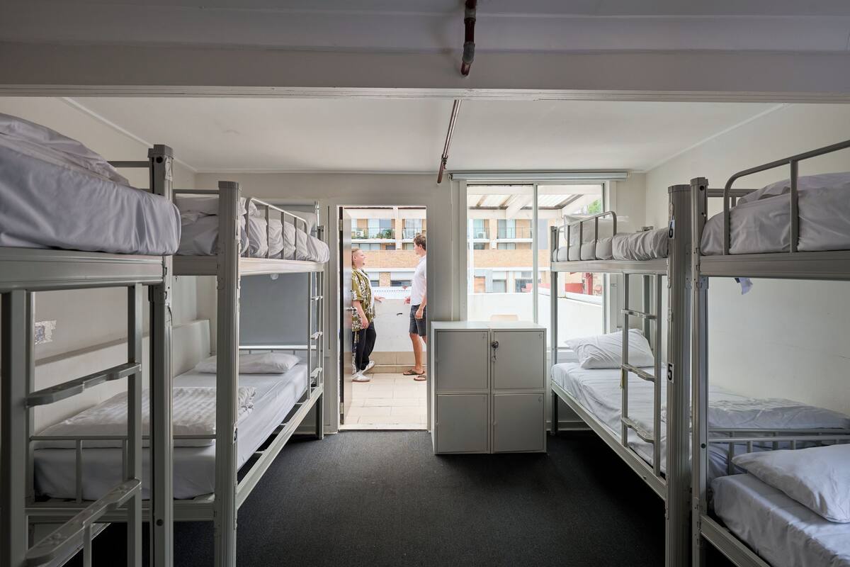 A dormitory room with bunk beds, a small cabinet, and a window providing a view of the outside.